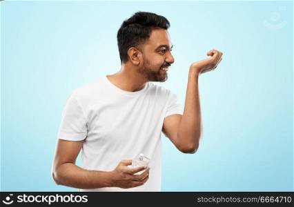 male perfumery, grooming and people concept - happy smiling young indian man smelling perfume over blue background. happy indian man with perfume over blue background