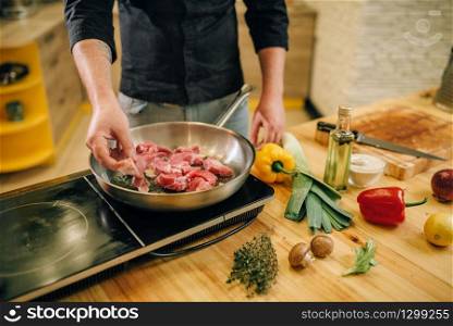 Male person cooking meat with herbs and seasonings in a pan on the kitchen. Man preparing boiled pork on table electric stove