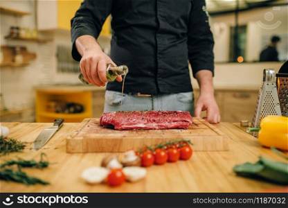 Male person hands seasoning piece of raw meat on wooden board, kitchen interior on background. Chef cooking tenderloin with vegetables, spices and herbs. Male person hands seasoning piece of raw meat