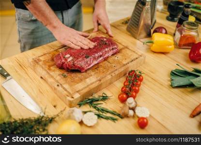 Male person hands seasoning piece of raw meat on wooden board, kitchen interior on background. Chef cooking tenderloin with vegetables, spices and herbs