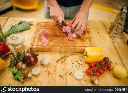 Male person hands with knife cuts raw meat into slices, top view, kitchen interior on background. Chef cooking tenderloin with vegetables, spices and herbs. Male person cuts raw meat into slices, top view