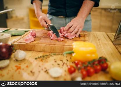 Male person hands with knife cuts raw meat into slices, top view, kitchen interior on background. Chef cooking tenderloin with vegetables. Male person cuts raw meat into slices, top view