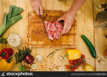 Male person hands with knife cuts raw meat into slices, top view, kitchen interior on background. Chef cooking tenderloin with vegetables, spices and herbs
