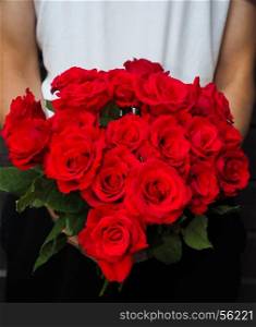 Male person holding a beautiful bouquet of red roses wearing white shirt and black pants against a gray wooden wall