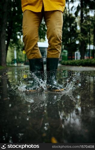 Male person in rain cape and rubber boots jumping in puddles, wet weather in alley. Man poses in summer park, rainy day. Water protection, drops. Male person in rubber boots jumping in puddles