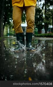 Male person in rain cape and rubber boots jumping in puddles, wet weather in alley. Man poses in summer park, rainy day. Water protection, drops. Male person in rubber boots jumping in puddles