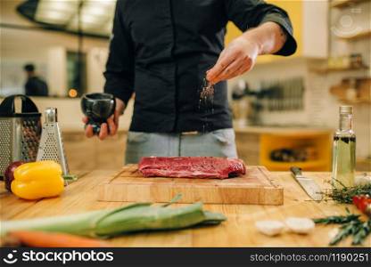 Male person marinating raw meat on wooden board, kitchen interior on background. Chef cooking tenderloin with vegetables, spices and herbs. Male person marinating raw meat on wooden board