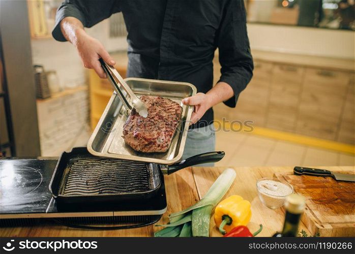 Male person puts the roasted meat into the pan on the kitchen. Man preparing boiled pork on table electric stove. Chef cooking tenderloin on countertop