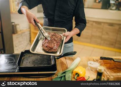 Male person puts the roasted meat into the pan on the kitchen. Man preparing boiled pork on table electric stove. Chef cooking tenderloin on countertop