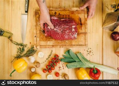 Male person seasoning piece of raw meat on wooden table, top view, kitchen interior on background. Chef cooking tenderloin with vegetables, spices and herbs
