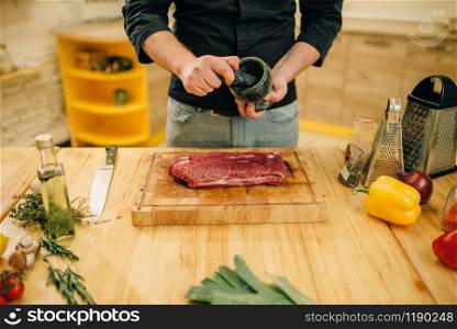Male person sprinkles raw meat with seasonings, kitchen interior on background. Chef cooking tenderloin with vegetables, spices and herbs. Male person sprinkles raw meat with seasonings