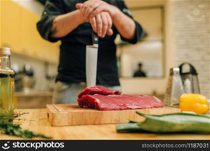 Male person with knife and piece of raw meat on wooden board, kitchen interior on background. Chef cooking tenderloin with vegetables, spices and herbs