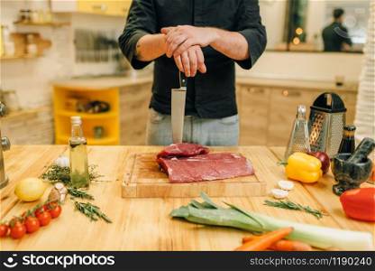 Male person with knife and piece of raw meat on wooden board, kitchen interior on background. Chef cooking tenderloin with vegetables, spices and herbs. Male person with knife and piece of raw meat