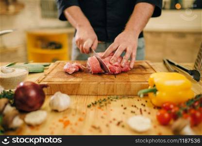 Male person with knife cuts raw meat into slices, closeup, kitchen interior on background. Chef cooking tenderloin with vegetables, spices and herbs