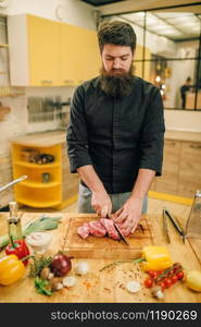 Male person with knife cuts raw meat into slices, closeup, kitchen interior on background. Chef cooking tenderloin with vegetables, spices and herbs. Male person with knife cuts raw meat into slices