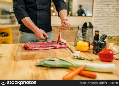 Male person with knife cuts raw meat on wooden board, kitchen interior on background. Chef cooking tenderloin with vegetables, spices and herbs