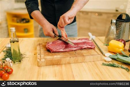 Male person with knife cuts raw meat on wooden board, kitchen interior on background. Chef cooking tenderloin with vegetables, spices and herbs. Male person with knife cuts raw meat on board