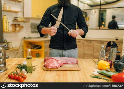 Male person with knife prepares to cut raw meat, kitchen interior on background. Chef cooking tenderloin with vegetables, spices and herbs
