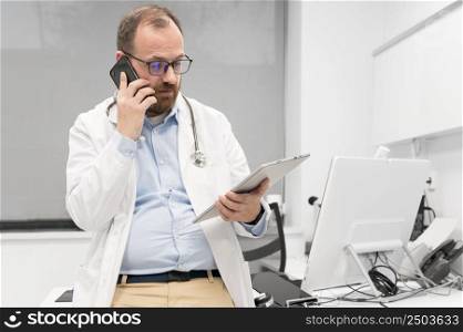 Male physician with stethoscope sitting at desk, talking on mobile phone and looking at digital tablet, discussing a patient diagnostic. High quality photography. Male physician with stethoscope sitting at desk, talking on mobile phone and looking at digital tablet, discussing a patient diagnostic.