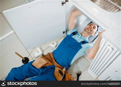 Male plumber in uniform installing drain pipe in the kitchen, top view. Handywoman with toolbag repair sink, sanitary equipment service at home. Plumber in uniform installing drain pipe, top view