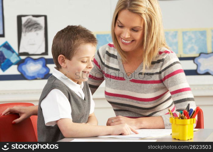 Male Primary School Pupil And Teacher Working At Desk In Classroom