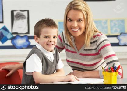 Male Primary School Pupil And Teacher Working At Desk In Classroom