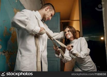 Male psychiatrist and crazy female patient in a fit of rage, mental hospital. Woman in strait jacket undergoing treatment in clinic for the mentally ill. Psychiatrist and female patient in a fit of rage