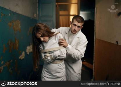 Male psychiatrist leads crazy female patient in straitjacket, mental hospital. Woman in strait jacket undergoing treatment in clinic for the mentally ill