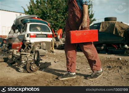 Male repairman holds toolbox on car scrapyard. Auto scrap, vehicle junk, automobile garbage. Abandoned, damaged and crushed transport, junkyard