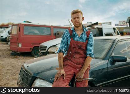 Male repairman poses on car junkyard. Auto scrap, vehicle junk, automobile garbage, abandoned, damaged and crushed transport. Male repairman poses on car junkyard