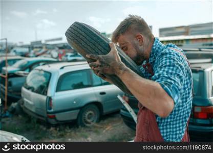 Male repairman pumps the tire with his mouth on car scrapyard. Auto scrap, vehicle junk, automobile garbage. Abandoned, damaged and crushed transport, junkyard