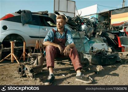 Male repairman sitting on the ground on car junkyard. Auto scrap, vehicle junk, damaged and crushed transport. Male repairman sitting on the ground, car junkyard
