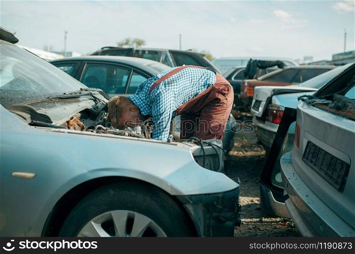 Male repairman works on car scrapyard. Auto scrap, vehicle junk, automobile garbage, abandoned, damaged and crushed transport