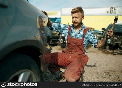 Male repairman works with hammer on car junkyard. Auto scrap, vehicle junk, automobile garbage, abandoned, damaged and crushed transport