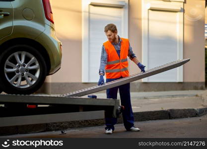 Male road worker in uniform preparing tow truck platform for car loading on city street side. Worker preparing tow truck platform for car