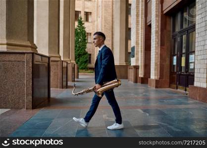 Male saxophonist holds instrument on the street, young talent. Musician with sax outdoors, musical performance in city, jazz performer. Male saxophonist holds instrument on the street