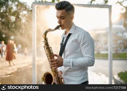 Male saxophonist plays melody on golden saxophone in summer park. Young musician with sax outdoors, musical performance in nature, jazz performer. Male saxophonist plays melody on saxophone in park