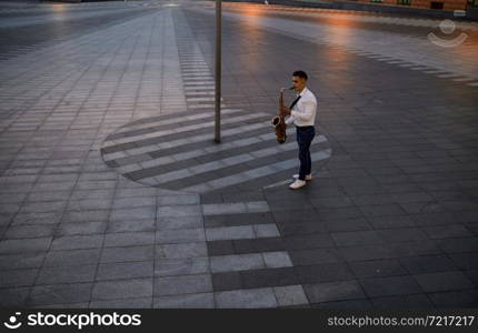 Male saxophonist plays the sax on the street in the evening, young talent. Musician with saxophone outdoors, musical performance in city, jazz performer. Saxophonist plays the sax on street in the evening