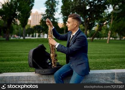 Male saxophonist takes the saxophone from the case on the alley in summer park. Musician with sax outdoors, musical performance in nature, jazz melody performer. Male saxophonist takes the saxophone from the case