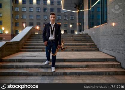 Male saxophonist with sax on the street in the evening, young talent. Musician with saxophone outdoors, musical performance in city, jazz performer. Male saxophonist with sax on street in the evening
