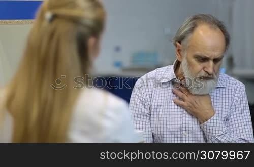 Male senior patient with beard visiting a doctor at the medical office. Elderly ill patient coughing and explaining his symptoms to female physician during checkup in doctor&acute;s office.
