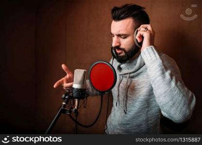 Male singer recording a song in music studio. Vocalist in headphones against microphone. Audio recording. Professional digital sound technologies. Male singer recording a song in music studio