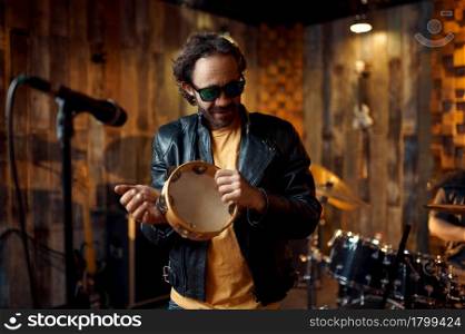 Male singer with tambourine at microphone, music performing on stage. Rock band performance or repetition in garage, man with musical instrument, live sound performers. Male singer with tambourine at microphone on stage