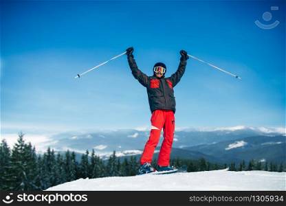 Male skier hands up on the top of mountain, blue sky, forest and snowy mountains on background. Winter active sport, extreme lifestyle. Downhill skiing