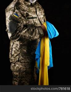 male soldier dressed in a military uniform of the Ukrainian army, he stands in his hand a yellow-blue state flag, dark background