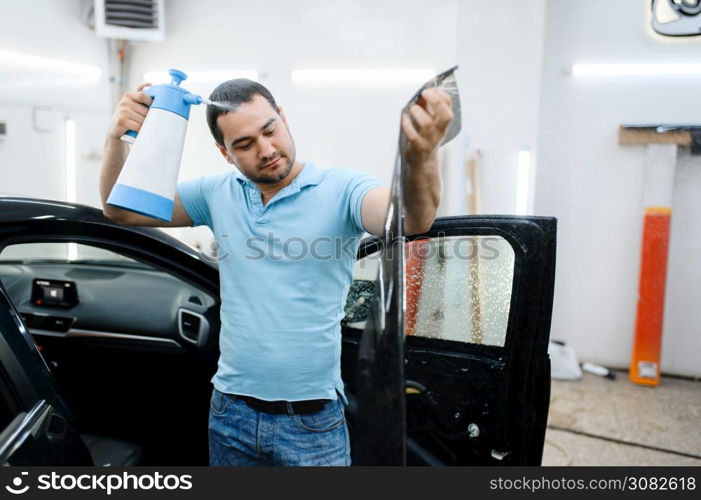 Male specialist with spray wetting car tinting, tuning service. Mechanic applying vinyl tint on vehicle window in garage, tinted automobile glass. Male specialist with spray wetting car tinting