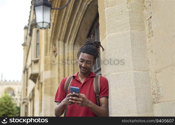 Male Student Checking Messages Or Social Media On Mobile Phone Outside University Building In Oxford UK