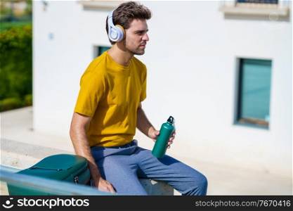Male student sitting outside using an aluminum water bottle, headphones and backpack.. Male sitting outside using an aluminum water bottle, headphones and backpack.