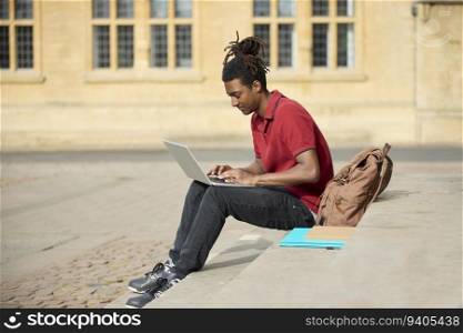 Male Student Working On Laptop Sitting On StepsOutside University Building In Oxford UK