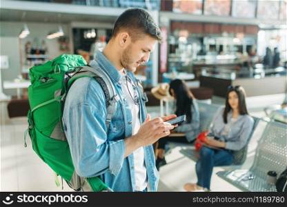 Male tourist with backpack holds mobile phone, female travelers waiting for departure in airport on background. Passengers with baggage in air terminal, happy journey, summer travel